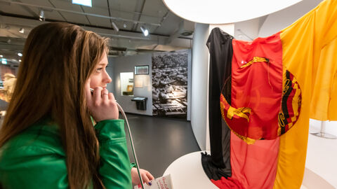 Eine Schülerin benutzt den Audio-Guide in der Dauerausstellung und steht dabei vor einer verblassenden Flagge der 
